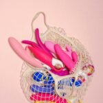 A variety of bright sex toys in a mesh bag on a soft pink background, showcasing modern sensuality.