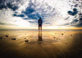 A lone figure stands on a tranquil beach during a vivid sunset, reflecting on the wet sand.