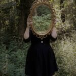 A woman holds an ornate mirror in a forest setting, reflecting nature.