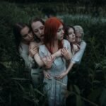 Four women embracing in a moody outdoor setting surrounded by lush greenery.