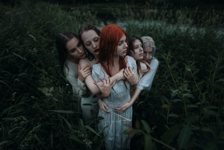 Four women embracing in a moody outdoor setting surrounded by lush greenery.